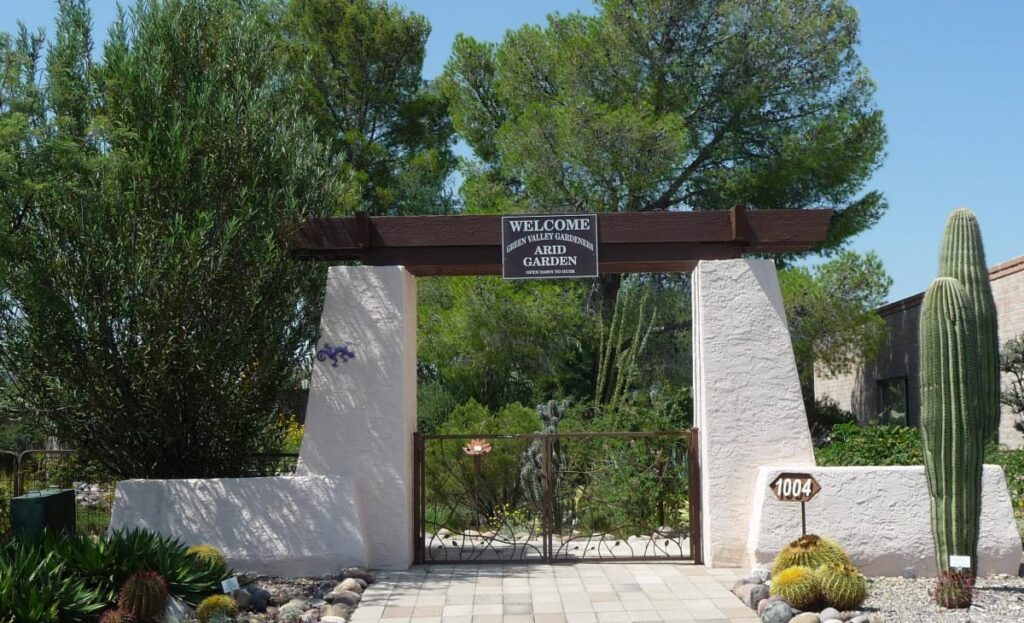 an archway of wood on stuccoed pillars with a leafy wrought iron gate, welcomes visitors to the Arid Garden on behalf of the Green Valley Gardeners